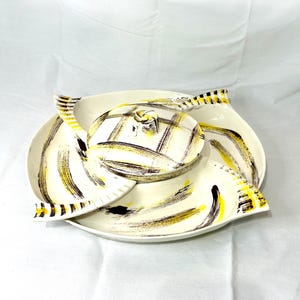 May include: A vintage serving set with a cream-colored base and a lid. The set features a unique design with black and yellow brushstroke patterns. The lid has a knob and a checkered pattern. The set includes a large platter and two smaller side dishes.