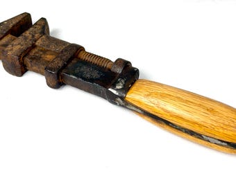 Puede incluir: Una llave inglesa ajustable antigua con mango de madera. Los componentes metálicos están muy oxidados, mostrando signos de edad y uso. El mango es de madera clara, contrastando con el metal oscuro. La llave está diseñada para apretar o aflojar pernos y tuercas.