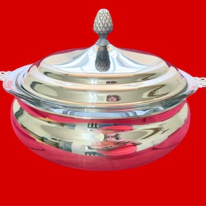 May include: A silver-colored serving dish with a lid, set against a red background. The dish has a rounded base and a lid topped with a decorative finial. The edges of the dish feature ornate detailing.