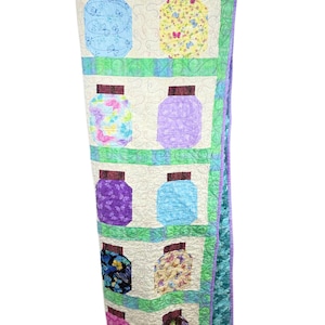 May include: A quilted wall hanging featuring a grid of ten jar-shaped fabric appliques on a cream background. Each jar has a dark brown lid and displays various colorful butterfly patterns in blue, yellow, purple, and black, separated by green borders.