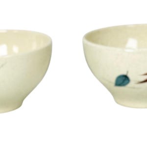 May include: Two off-white ceramic cups with a speckled finish. The left cup features a single yellow leaf illustration. The right cup displays a blue leaf and a larger brown oak leaf design. Both cups have a rounded bowl shape and a side handle.