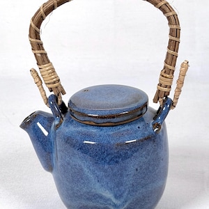 May include: A small, blue ceramic teapot with a woven handle and lid. The teapot has a rounded body, a spout, and a lid. The handle is made of woven brown and tan material. The teapot is a shade of blue with darker blue accents.