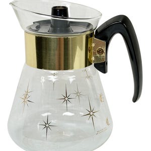 May include: A clear glass coffee carafe featuring a gold-toned metal band around the neck and a black plastic handle. The glass body is decorated with gold and black starburst patterns. A black plastic lid is fitted to the top of the carafe.