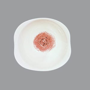 May include: A white ceramic bowl with a slightly irregular, rounded shape. The interior of the bowl features a centered, circular design in a textured, terracotta-colored pigment that resembles a hand-drawn or stamped spiral pattern.