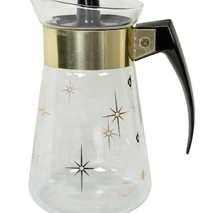 May include: A clear glass coffee carafe featuring a gold-toned metal band around the neck and a black plastic handle. The glass body is decorated with gold starburst patterns and includes small black numerical markings for measuring liquid capacity.