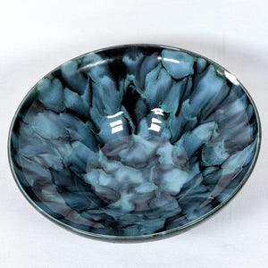 May include: A round ceramic bowl with a glossy finish. The bowl features a unique pattern of blue and teal hues, creating a marbled effect. The rim of the bowl is a darker shade of blue.