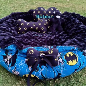 May include: A round pet bed with a blue Batman-themed design. The bed features a plush, dark blue interior and a blue exterior with Batman graphics. Includes a bone-shaped pillow and a bow.