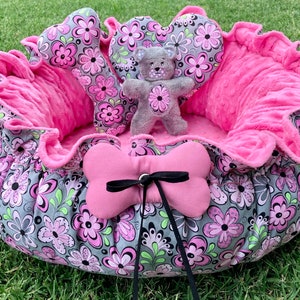 May include: A pink and gray dog bed with a floral pattern. The bed has a plush pink lining and comes with a gray teddy bear toy, a pink bone-shaped pillow, and a pink pillow with a black bow.