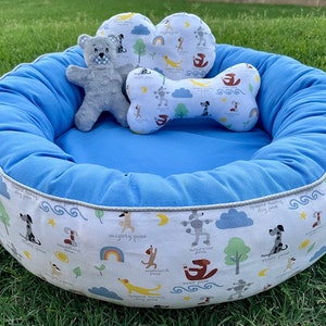May include: A blue and white dog bed with a plush gray teddy bear and a bone-shaped pillow. The bed has a white background with a pattern of dogs and clouds. The text on the bed reads "speed racing dog pose", "mighty pose", "warrior pose", and "ground training dog pose".