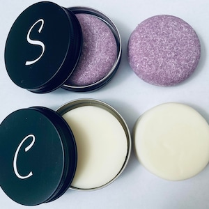 Puede incluir: Dos latas redondas negras con letras blancas, una con la letra "S" y la otra con la letra "C". Cada una contiene una pastilla de champ&uacute; s&oacute;lido. Una es de color lavanda y la otra es blanca. Tambi&eacute;n se ven dos pastillas de champ&uacute; s&oacute;lido adicionales.