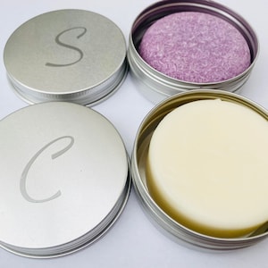 May include: Two silver tins with lids, one containing a white solid bar of soap and the other containing a purple solid bar of soap. The lids have the letters "S" and "C" engraved on them.