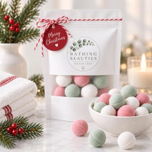 以下が含まれることがあります： Bathing Beauties Eco Bath & Bodyとラベルが貼られた白いスタンドパウチ。中には白、ピンク、セージグリーンの丸いバスボムが入っています。Merry Christmasと書かれた赤い円形のタグが、赤と白の紐でパウチの上部に結び付けられています。