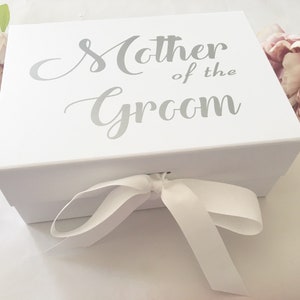 May include: White gift box with a white satin ribbon tied in a bow. The box has the words "Mother of the Groom" printed in silver on the top.