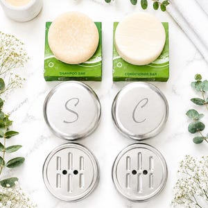 Könnte beinhalten: Ein Set aus zwei runden Shampoo- und Conditioner-Stücken in grünen Schachteln, zwei silbernen Metalldosen mit den eingravierten Buchstaben S und C auf den Deckeln sowie zwei passenden silbernen Metallseifenschalen mit Ablaufrillen.