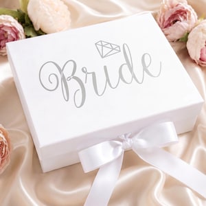 May include: A white rectangular gift box featuring the word Bride in elegant silver script lettering with a small diamond icon above the text. The box is secured with a white satin ribbon tied in a bow at the front center, creating a clean look.