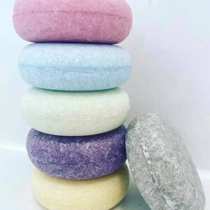 Puede incluir: Una pila de seis champ&uacute;s s&oacute;lidos redondos en colores pastel: rosa, azul, verde, morado, amarillo y gris. Las barras tienen una superficie texturizada, lo que sugiere una f&oacute;rmula concentrada para el cuidado del cabello.