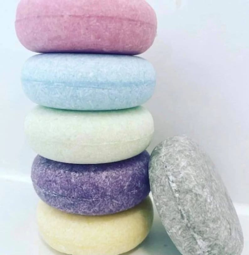 Puede incluir: Una pila de seis champ&uacute;s s&oacute;lidos redondos en colores pastel: rosa, azul, verde, morado, amarillo y gris. Las barras tienen una superficie texturizada, lo que sugiere una f&oacute;rmula concentrada para el cuidado del cabello.