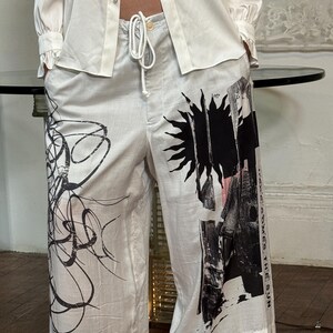 Puede incluir: Pantalones blancos de pernera ancha con un dise&ntilde;o abstracto negro y el texto "HERE COMES THE SUN". Los pantalones tienen cintura con cord&oacute;n y se combinan con una camisa blanca abotonada.