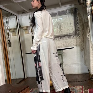 Puede incluir: Una persona con una blusa color crema y pantalones blancos de pernera ancha. Los pantalones presentan un dise&ntilde;o gr&aacute;fico en blanco y negro con texto. El conjunto se completa con botas negras de tac&oacute;n. El fondo incluye un espejo y una alfombra estampada.