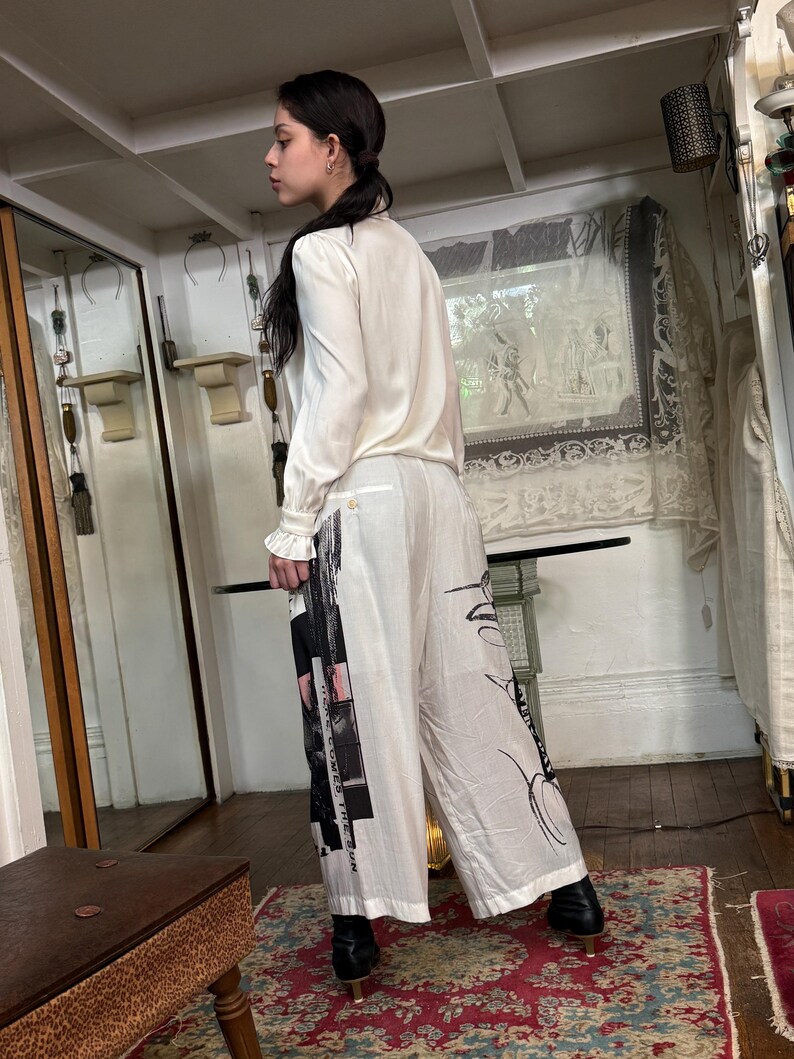 Puede incluir: Una persona con una blusa color crema y pantalones blancos de pernera ancha. Los pantalones presentan un dise&ntilde;o gr&aacute;fico en blanco y negro con texto. El conjunto se completa con botas negras de tac&oacute;n. El fondo incluye un espejo y una alfombra estampada.
