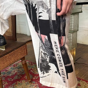 Puede incluir: Pantalones blancos con un dise&ntilde;o abstracto en negro y rosa. El texto "HERE COMES THE SUN" est&aacute; impreso verticalmente en el lateral. Los pantalones se combinan con zapatos negros.