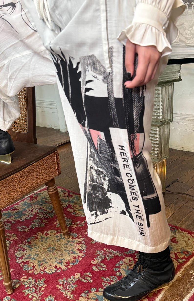 Puede incluir: Pantalones blancos con un dise&ntilde;o abstracto en negro y rosa. El texto "HERE COMES THE SUN" est&aacute; impreso verticalmente en el lateral. Los pantalones se combinan con zapatos negros.