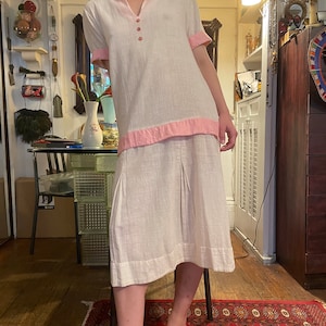 May include: A white linen dress with a pink trim around the collar and hem. The dress has a button-up collar and short sleeves.