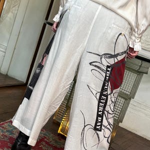 Puede incluir: Pantalones blancos de pernera ancha con dise&ntilde;os abstractos en negro y rojo y el texto "THE SUN EVERY DAY". Los pantalones tienen cintura alta y cierre de bot&oacute;n. Parte de una colecci&oacute;n de ropa.