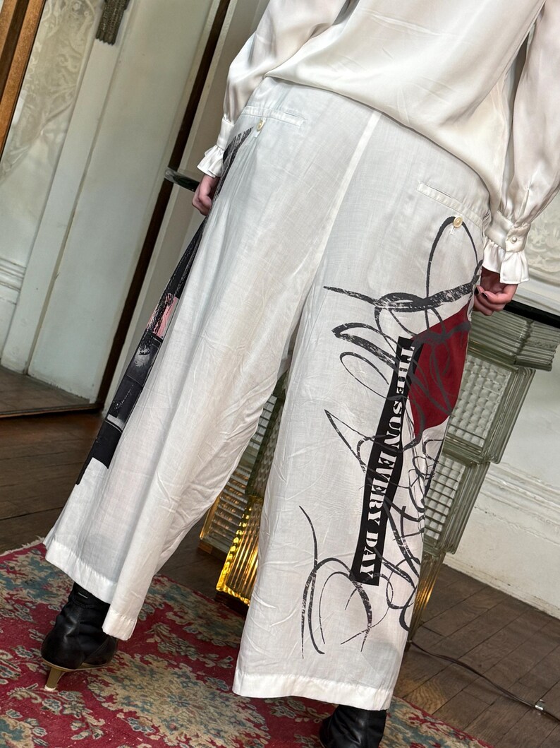 Puede incluir: Pantalones blancos de pernera ancha con dise&ntilde;os abstractos en negro y rojo y el texto "THE SUN EVERY DAY". Los pantalones tienen cintura alta y cierre de bot&oacute;n. Parte de una colecci&oacute;n de ropa.