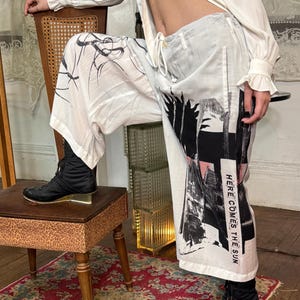 Puede incluir: Pantalones blancos con dise&ntilde;os abstractos en negro y rosa y el texto "HERE COMES THE SUN". Los pantalones tienen cintura con cord&oacute;n. Se usan botas negras con los pantalones. Los pantalones son parte de una colecci&oacute;n de moda.
