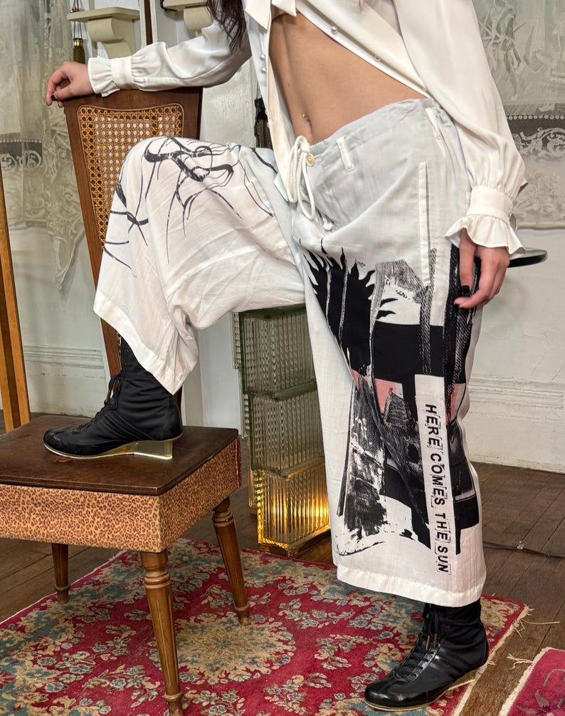 Puede incluir: Pantalones blancos con dise&ntilde;os abstractos en negro y rosa y el texto "HERE COMES THE SUN". Los pantalones tienen cintura con cord&oacute;n. Se usan botas negras con los pantalones. Los pantalones son parte de una colecci&oacute;n de moda.