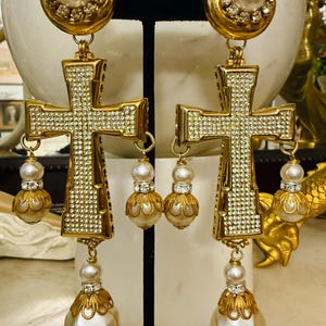 May include: A pair of gold-tone drop earrings featuring a large cross pendant encrusted with clear rhinestones. Each earring includes a circular top with a central faux pearl surrounded by rhinestones, and multiple dangling faux pearl accents.