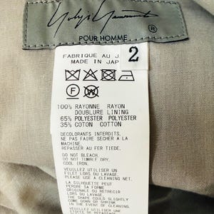 Puede incluir: Primer plano de una etiqueta de ropa con el texto "Yohji Yamamoto POUR HOMME" en escritura negra. La etiqueta incluye instrucciones de cuidado y detalles de la composici&oacute;n de la tela: 100% ray&oacute;n, 65% poli&eacute;ster y 35% algod&oacute;n. El n&uacute;mero "2" est&aacute; impreso en la etiqueta.