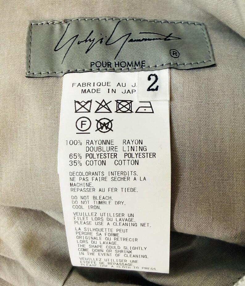 Puede incluir: Primer plano de una etiqueta de ropa con el texto "Yohji Yamamoto POUR HOMME" en escritura negra. La etiqueta incluye instrucciones de cuidado y detalles de la composici&oacute;n de la tela: 100% ray&oacute;n, 65% poli&eacute;ster y 35% algod&oacute;n. El n&uacute;mero "2" est&aacute; impreso en la etiqueta.