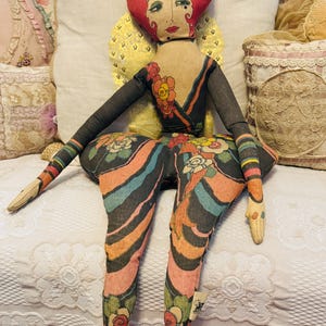 May include: A vintage cloth doll with a red cap and painted face. The doll wears a black outfit with colorful stripes and floral patterns. The doll's arms and legs are elongated, and the doll is seated on a white surface.
