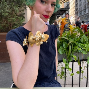 May include: A gold-toned bracelet with a cluster of white and gold beads and leaves. The bracelet is worn on the wrist of a person with green hair.