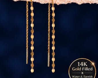 Puede incluir: Un par de pendientes de hilo rellenos de oro con una cadena fina y una fila vertical de pequeños discos dorados planos y circulares. Una insignia circular muestra el texto 14K Gold Filled Water & Tarnish Resistant.