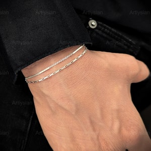 May include: Two silver bracelets are worn on a wrist. The top bracelet is a delicate chain, and the bottom bracelet features rectangular links. The bracelets are displayed against a black background.