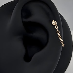 Könnte beinhalten: Ein goldfarbener Ohrschmuck mit einem kleinen Stecker und einer herabhängenden Kette aus verbundenen Kreisen. Der Ohrring wird auf einem schwarzen Ohrmodell vor weißem Hintergrund gezeigt.