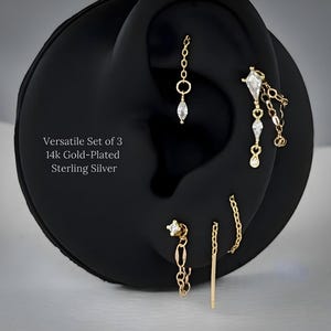 Pode incluir: Um conjunto de três brincos em prata esterlina banhados a ouro 14k, exibidos em um modelo de orelha preta. Os brincos apresentam vários designs, incluindo correntes, barras e detalhes de pedras preciosas. Texto: "Versatile Set of 3 14k Gold-Plated Sterling Silver."