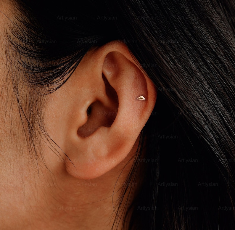 May include: A close-up of a person's ear with a small gold triangle stud earring in the lobe.