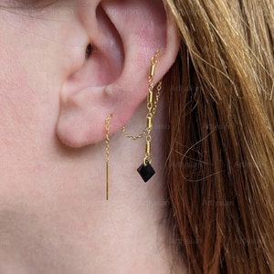May include: Gold-toned ear jewelry featuring a linear drop earring and a chain earring with a black diamond-shaped charm. The earrings are worn on the ear, showcasing their design and style. The jewelry is delicate and elegant.