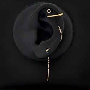 May include: Gold-coloured earrings displayed on a black ear-shaped form. The earrings include a small hoop, a chain earring, and a stud earring. The earrings are set against a black background.