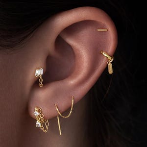 May include: Close-up of an ear adorned with multiple gold earrings. The earrings include a small bar, a chain with a dangling rectangular charm, a chain with a dangling rectangular gem, and a chain with a small round gem.