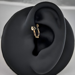 May include: A gold chain earring is displayed in an ear piercing. The earring features a small, flat disc at one end and a chain of rectangular links. The earring is set against a black background.