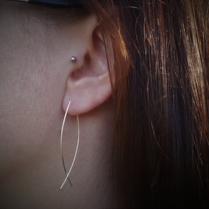 Può includere: Primo piano di un orecchio con un orecchino lineare curvo in argento. L'orecchino è in metallo sottile e lucido che si incrocia nella parte inferiore. La persona ha i capelli castani e un piccolo piercing d'argento.