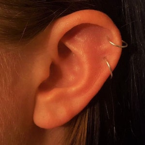 May include: A close-up of a person's ear with two silver hoop earrings. The earrings are in the upper lobe and helix piercings. The ear is a light orange color and the background is dark brown.