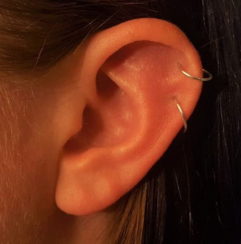 May include: A close-up of a person's ear with two silver hoop earrings. The earrings are in the upper lobe and helix piercings. The ear is a light orange color and the background is dark brown.