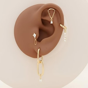 Puede incluir: Una colección de pendientes dorados exhibidos en una oreja. Los pendientes incluyen una cadena, un pendiente colgante en forma de diamante, un aro y una serie de pequeñas gemas colgantes. La oreja es de color marrón medio, sobre un fondo claro.