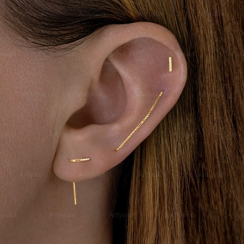 Op de afbeelding: Close-up van een oor versierd met meerdere gouden oorbellen. De oorbellen omvatten een kleine staafstud, een lange kettingachtige oorbel en een verticale staafoorbel. De sieraden zijn geplaatst tegen de achtergrond van de huid en het haar van een persoon.